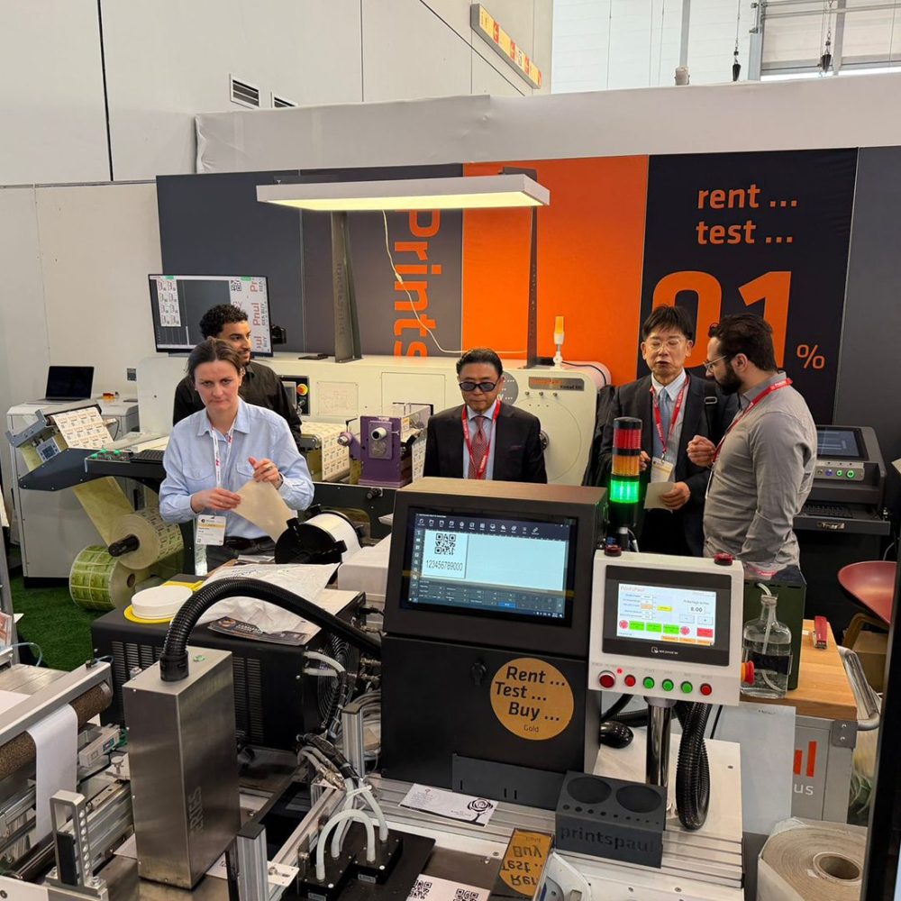 PrintsPaul standında makineler ve Rent-Test-Buy duvarı